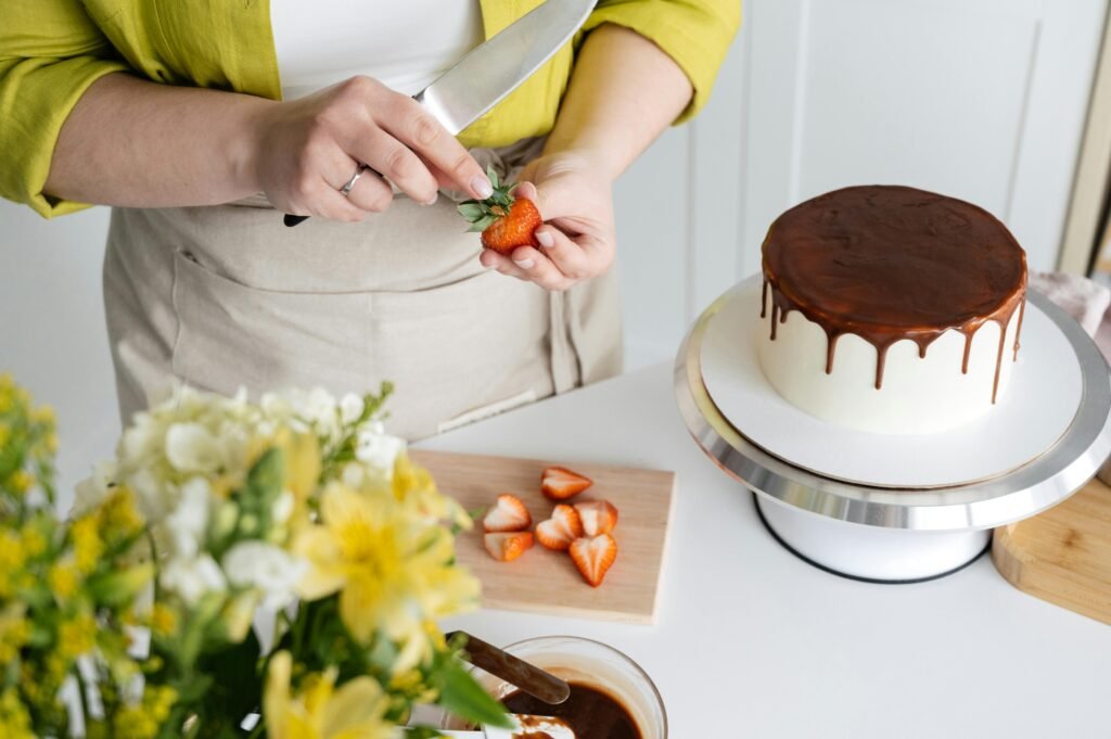Crop unrecognizable female wearing apron cutting fresh strawberries with knife on wooden board for decoration of cake with chocolate glaze
